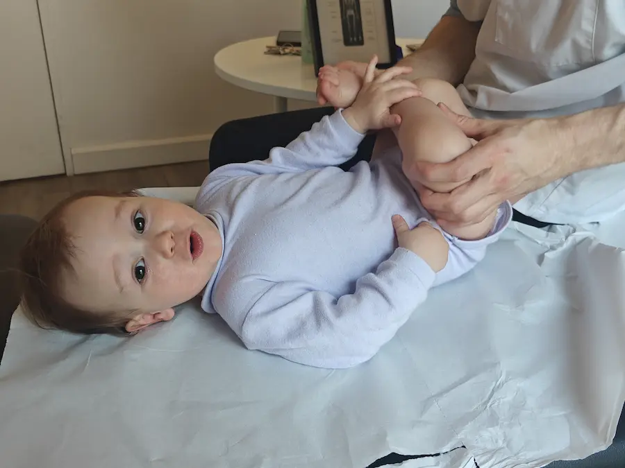 Gros plan d'un bébé en body bleu ciel sur une table d'examen. Les mains de l'ostéopathe Martin Ducuing, portant une blouse blanche, manipulent doucement les hanches et les jambes du nourrisson pour un test de mobilité articulaire.